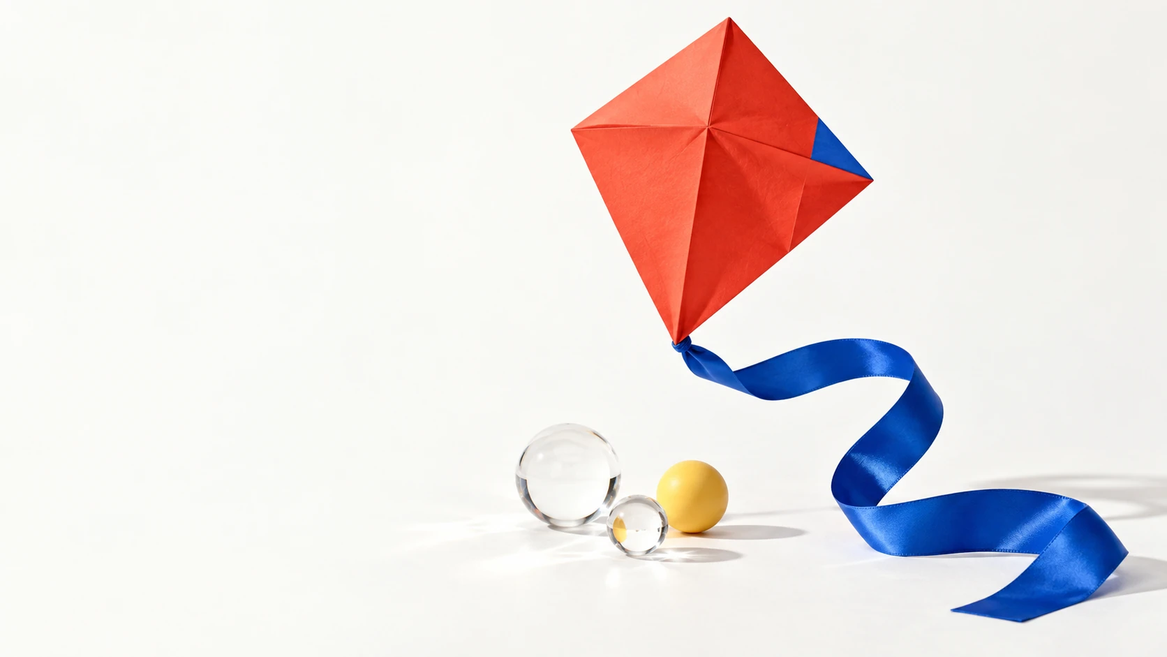 A red folded-paper kite with a flowing cobalt ribbon, glass spheres and a yellow ball.